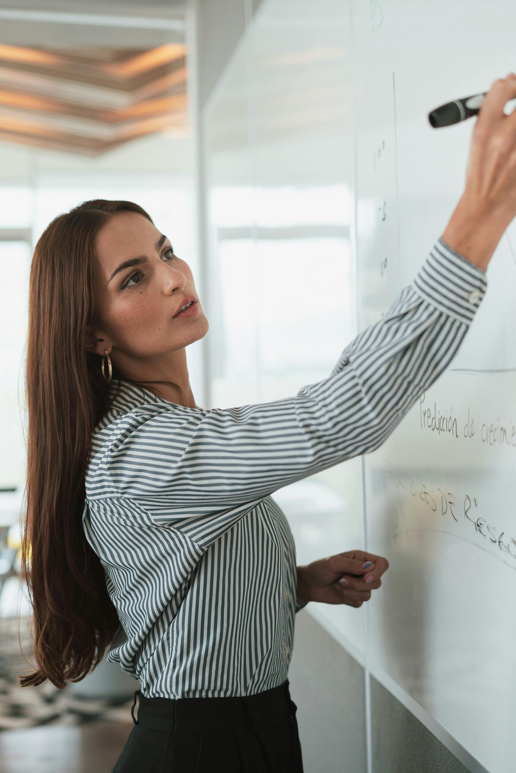 Woman in striped shirt presenting on a whiteboard in a modern office setting.